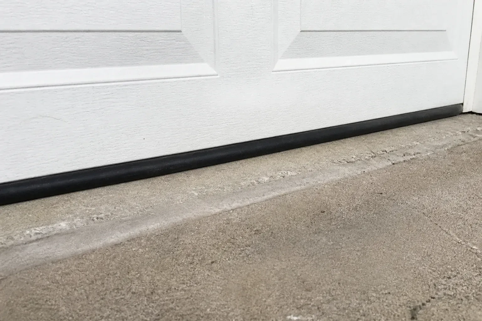 Black bottom seal along a closed white garage door at a concrete threshold. — Garage Door Bottom Seal Replacement, Vaughan