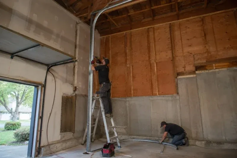 One person on a stepladder working beside a tall curved garage-door track while another kneels beside loose track sections in an unfinished garage. — High-Lift Garage Door Conversion, Vaughan